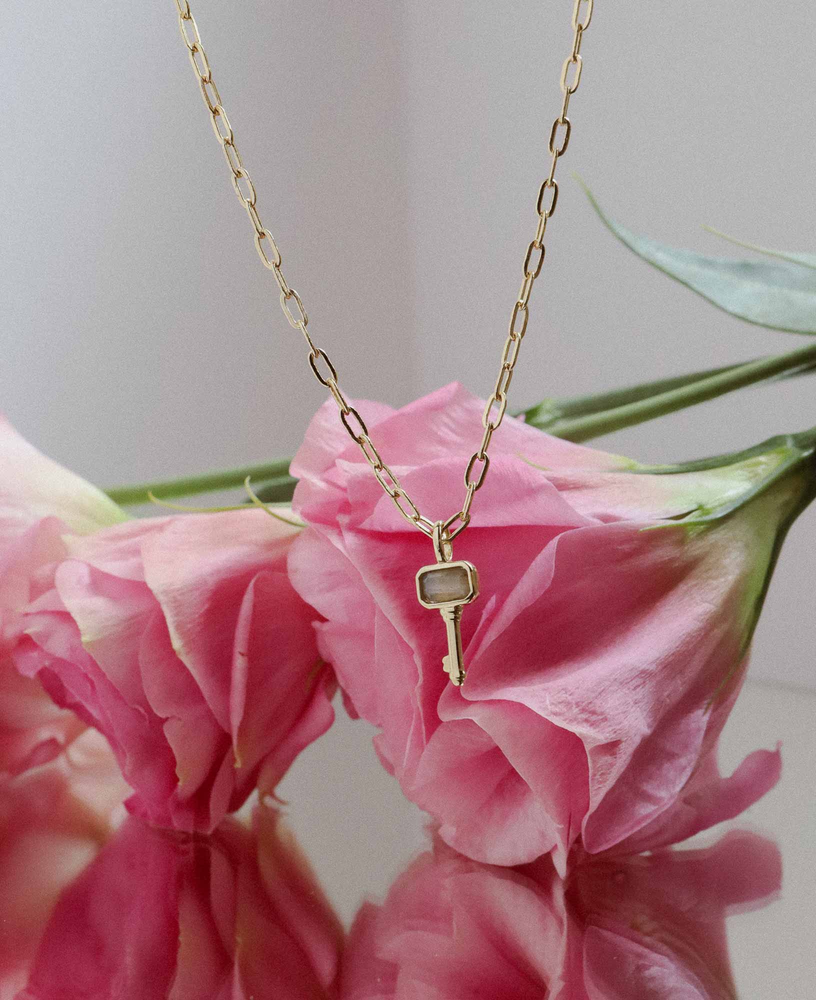 flat lay picture of the Francoise Labradorite Key Pendant Chain Necklace dangling in front of a bouquet of pink flowers.