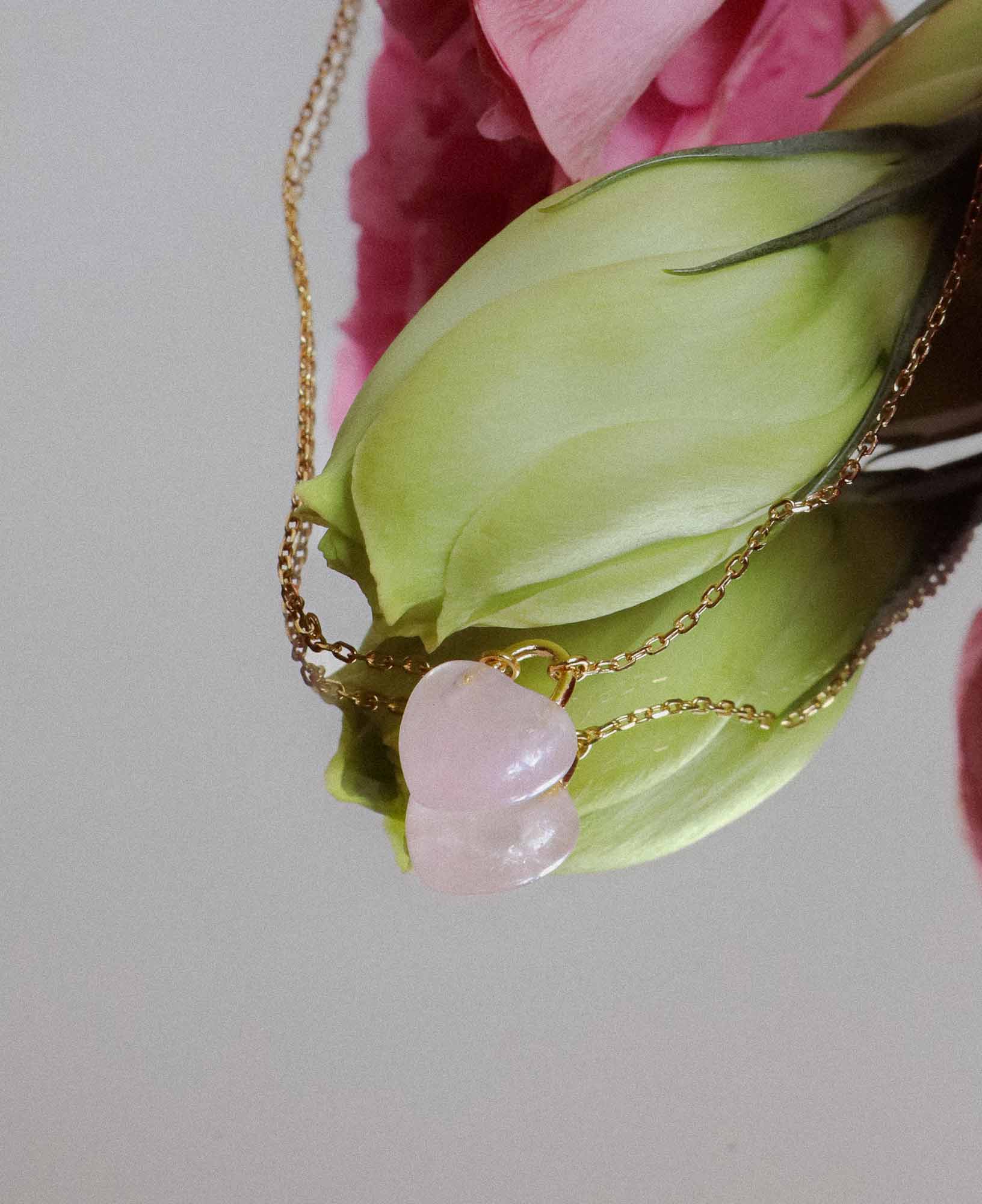 the Desiree Eternal Heart Rose Quartz Pendant Necklace placed on a mirror in front of a flower bud.