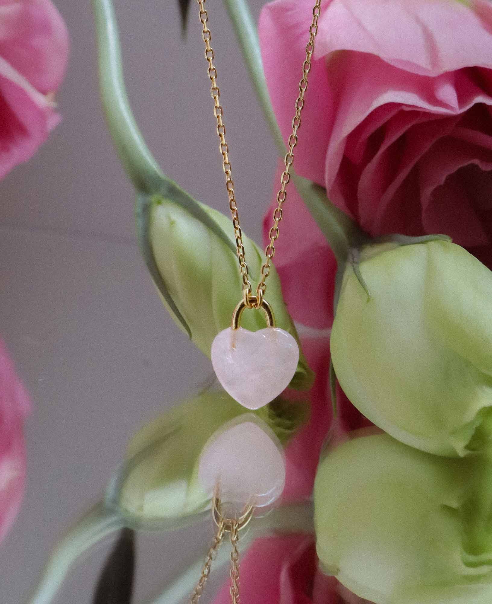 the Desiree Eternal Heart Rose Quartz Pendant Necklace dangling in front of a bouquet of pink flowers and flower buds.