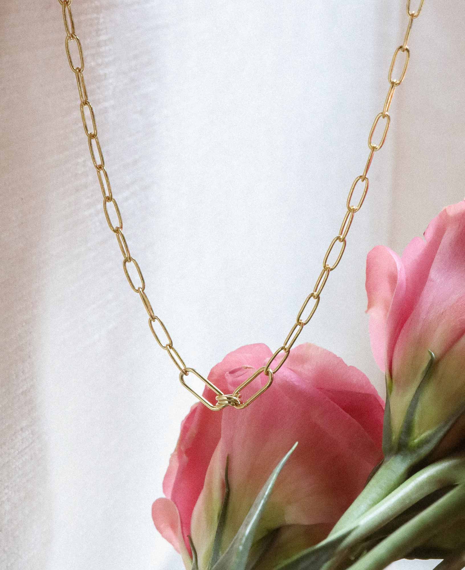 aesthetic flat lay shot of the Charlene Love Knot Gold Paperclip Chain Necklace dangling in front of some pink flowers.