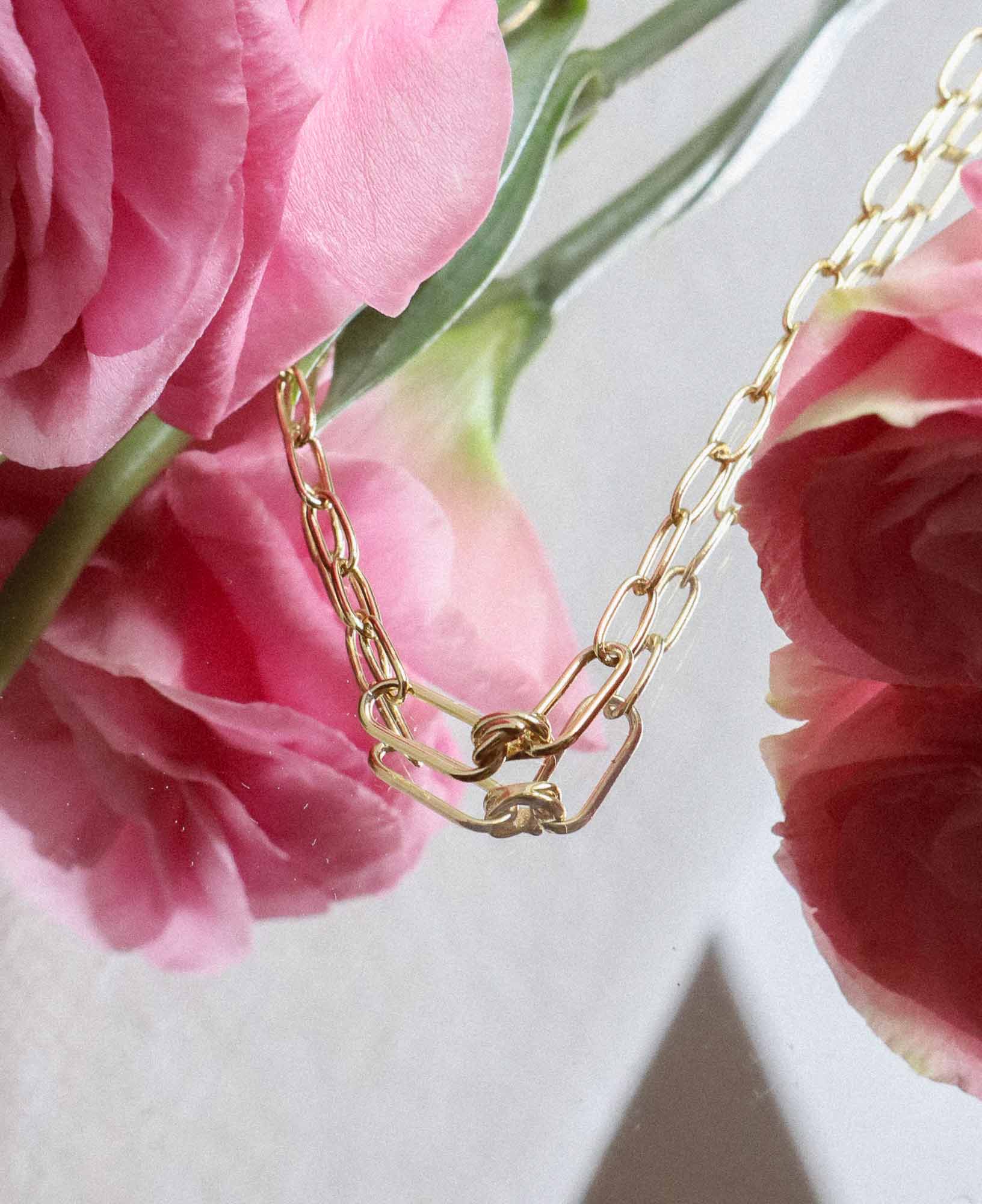 aesthetic flat lay shot of the Charlene Love Knot Gold Paperclip Chain Necklace around some pink flowers on placed on a mirror.