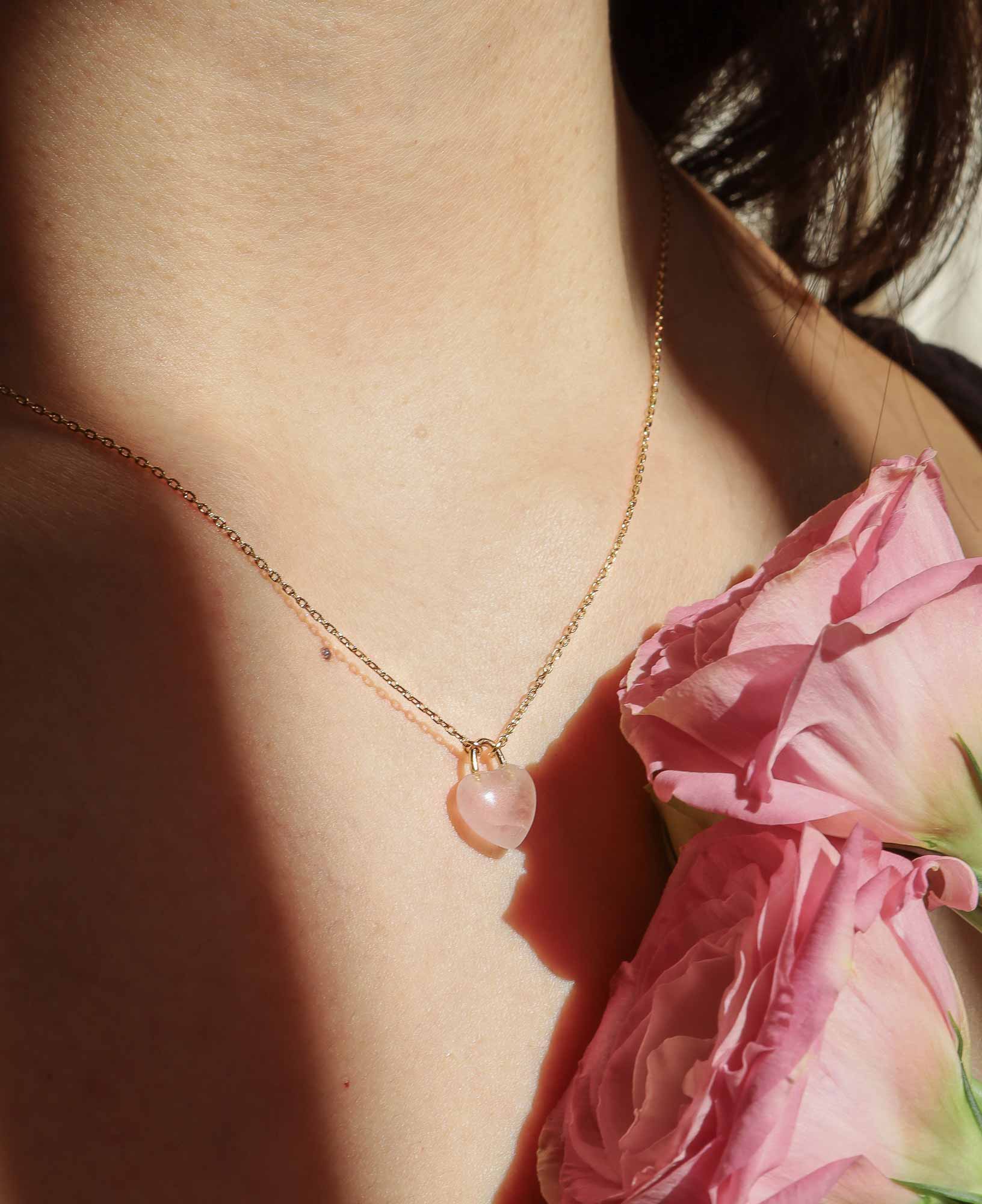 the Desiree Eternal Heart Rose Quartz Pendant Necklace featured on a model decorated with some seasonal pink flowers.