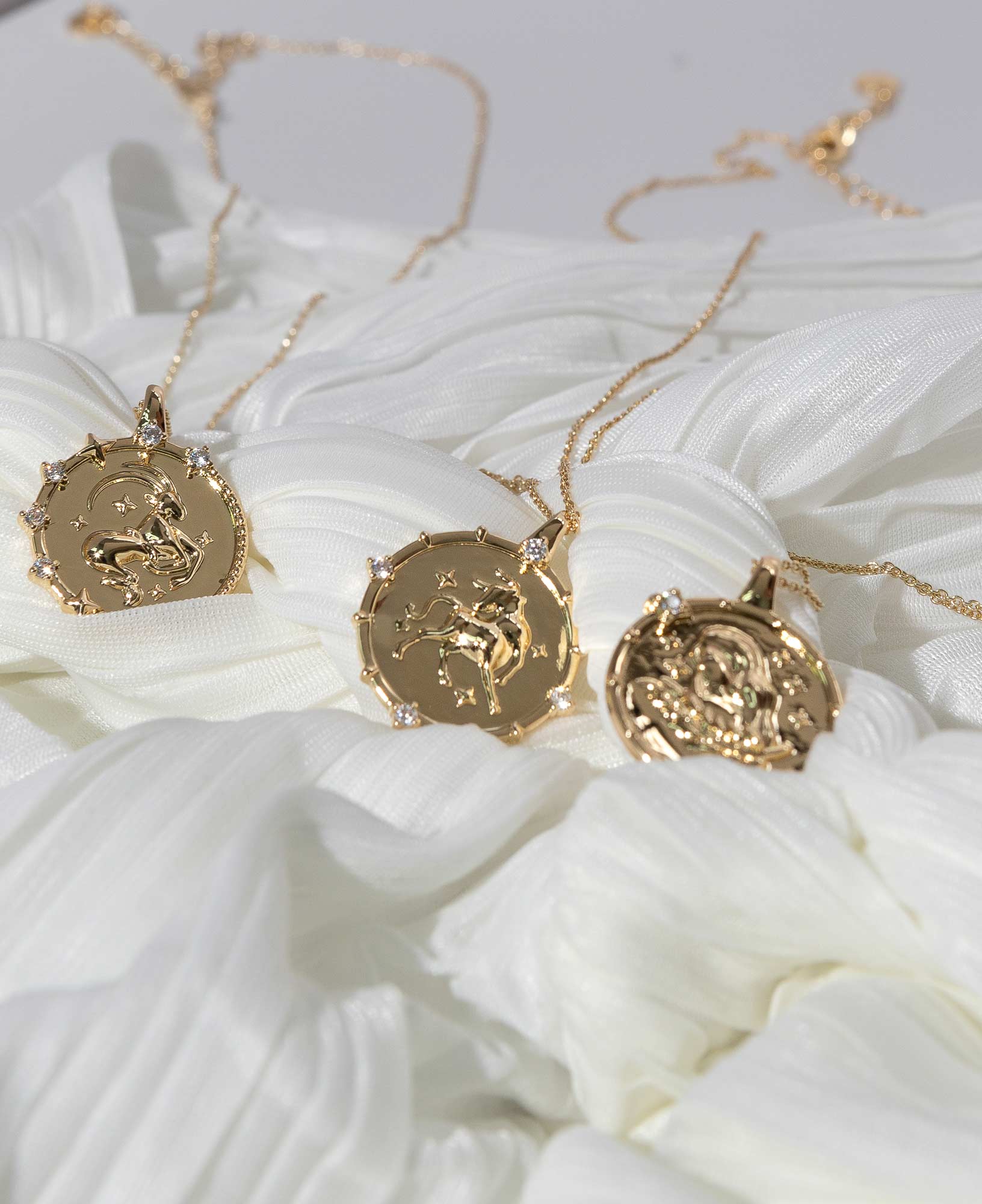 artistic close-up product picture of three Earth Signs' Horoscope Gold Pendant Necklaces laid on white chiffon fabric.