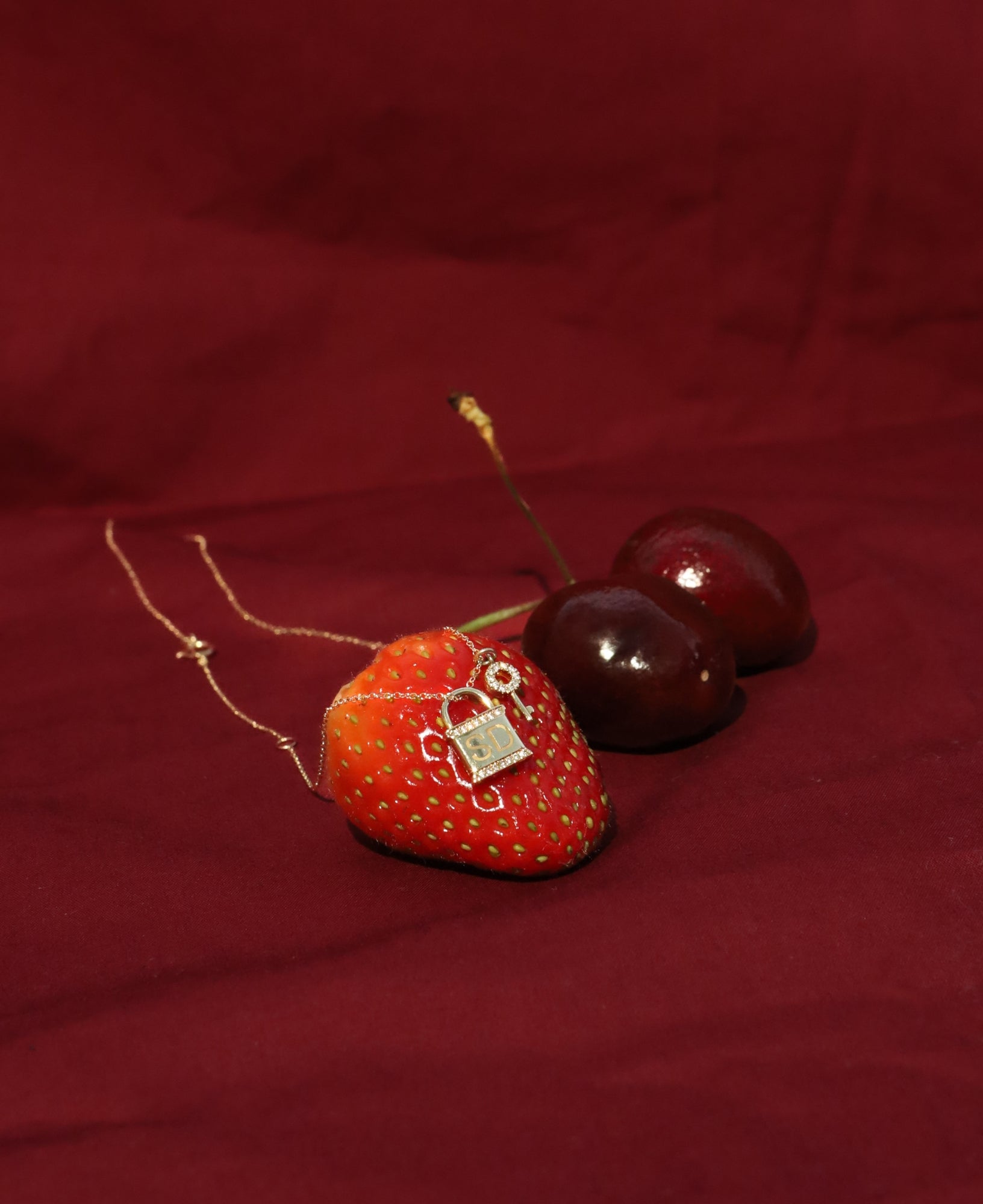 Strawberry with the leighton custom lock-and-key engraved diamond pendant necklace and two cherries on a red fabric background