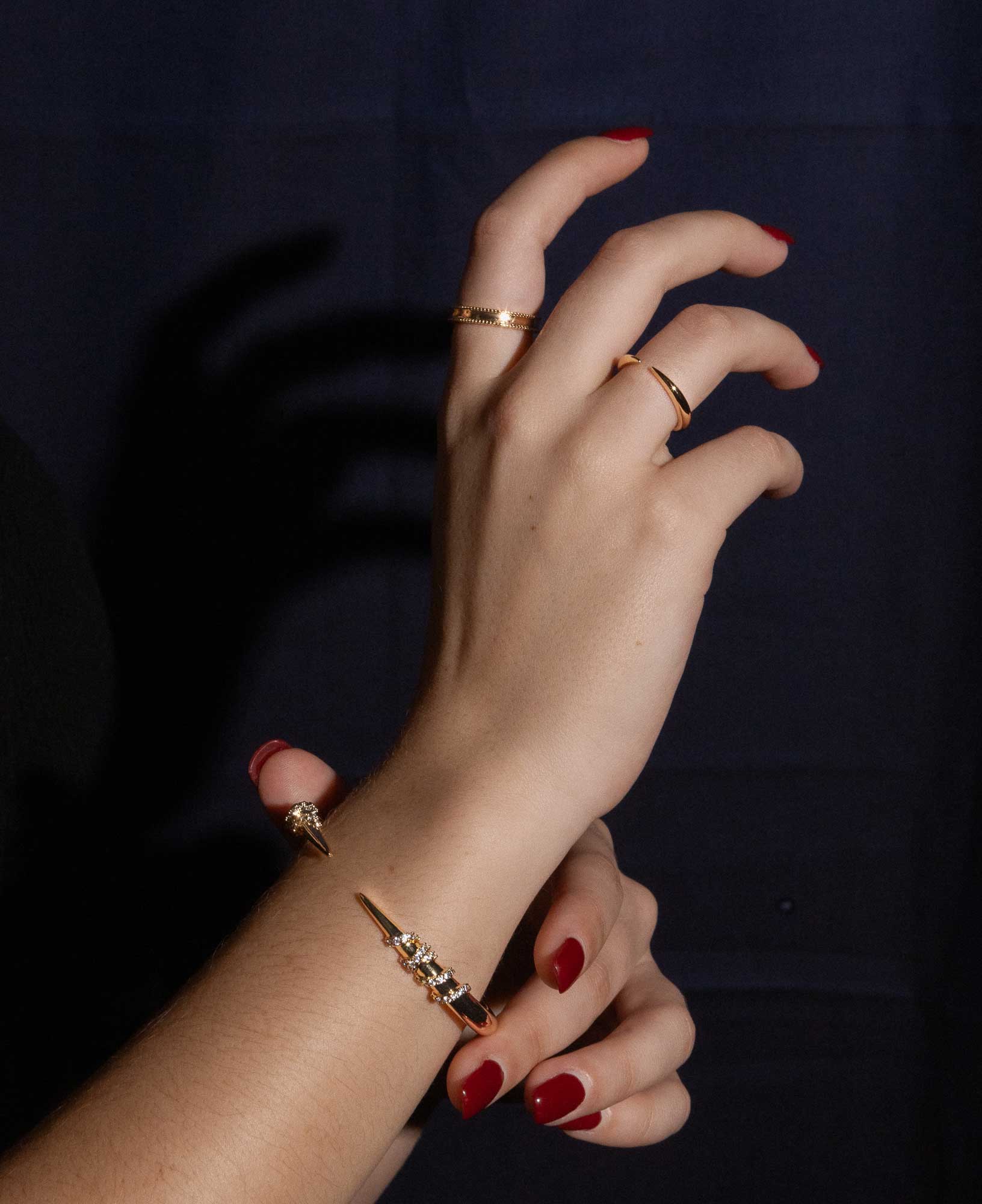 dark and festive close-up shot of model's hand featuring the Gladys Beaded Gold Band Ring, Imogen Gold Cuff Open Ring and Vesper CZ Cluster Claw Bangle.